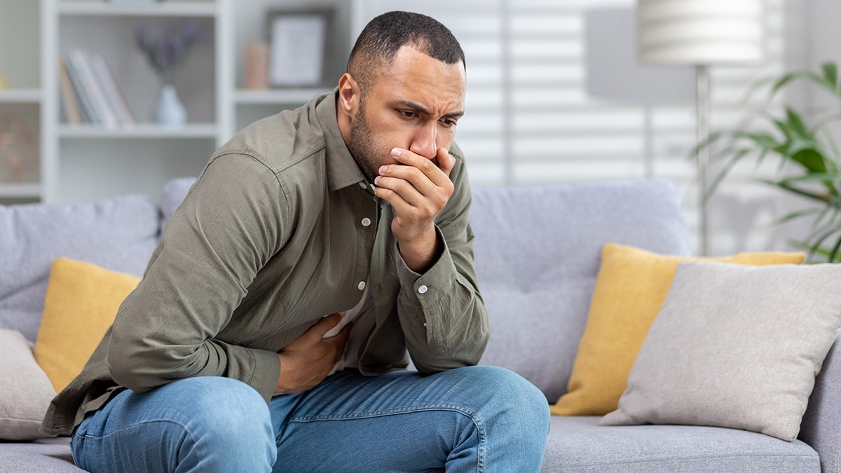 Man sitting on couch holds mouth looking sick, signaling stomach issues and vomiting.