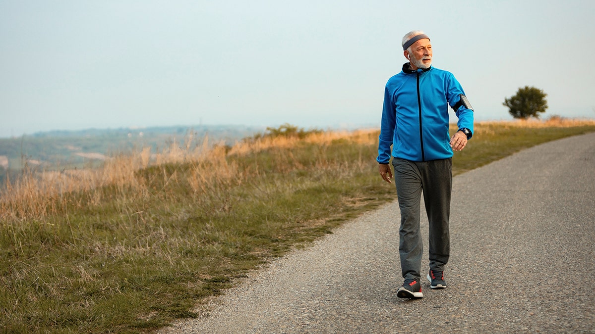 An older man walking along a rural path wearing a blue jacket, gray pants, and sneakers on a calm day.