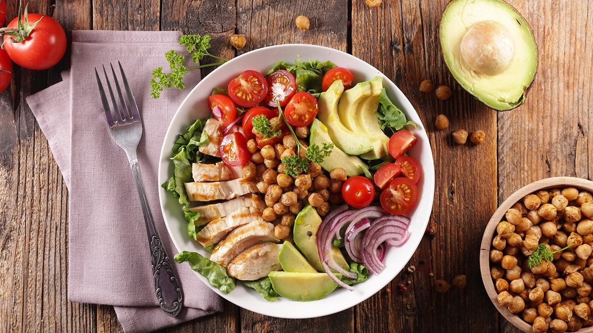 A healthy salad bowl with grilled chicken, avocado slices, cherry tomatoes, red onion, chickpeas, and lettuce on a rustic wooden table.