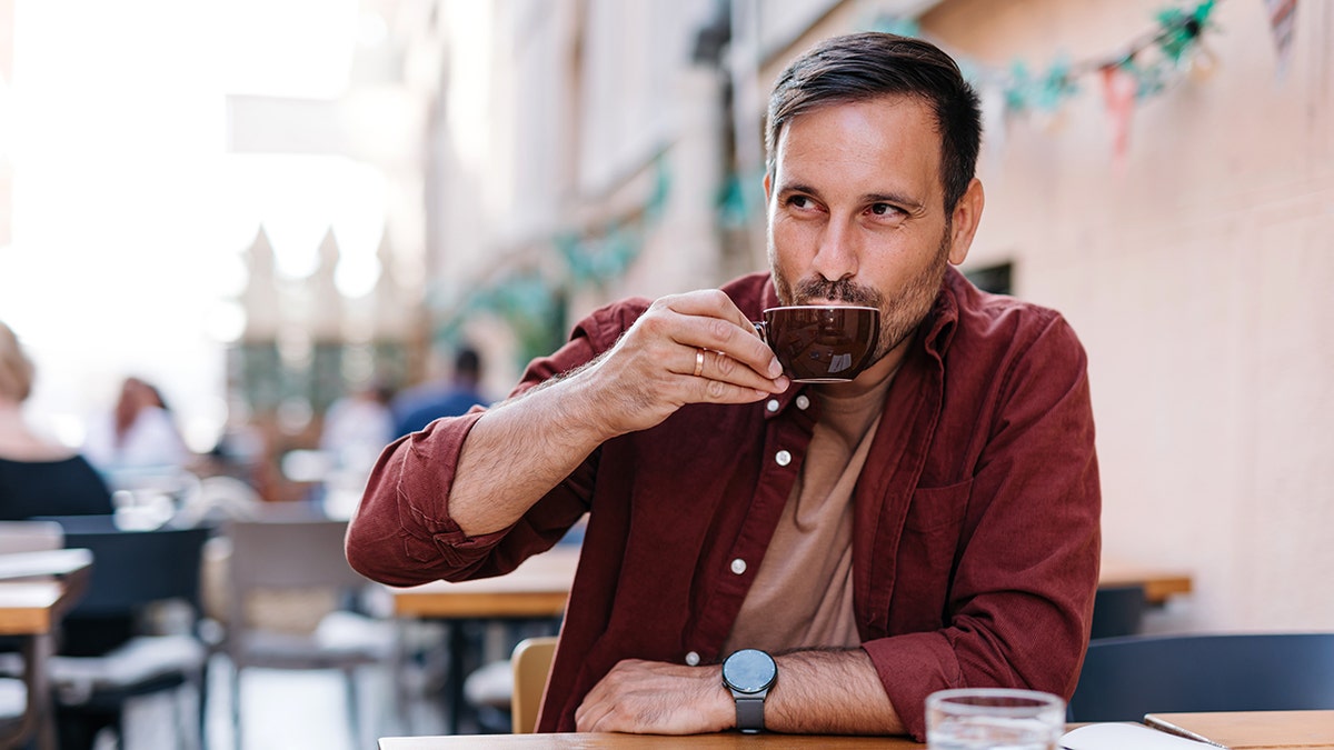A man in casual attire sips coffee while seated at a bustling outdoor cafe, capturing a serene yet lively energy in an engaging urban environment.