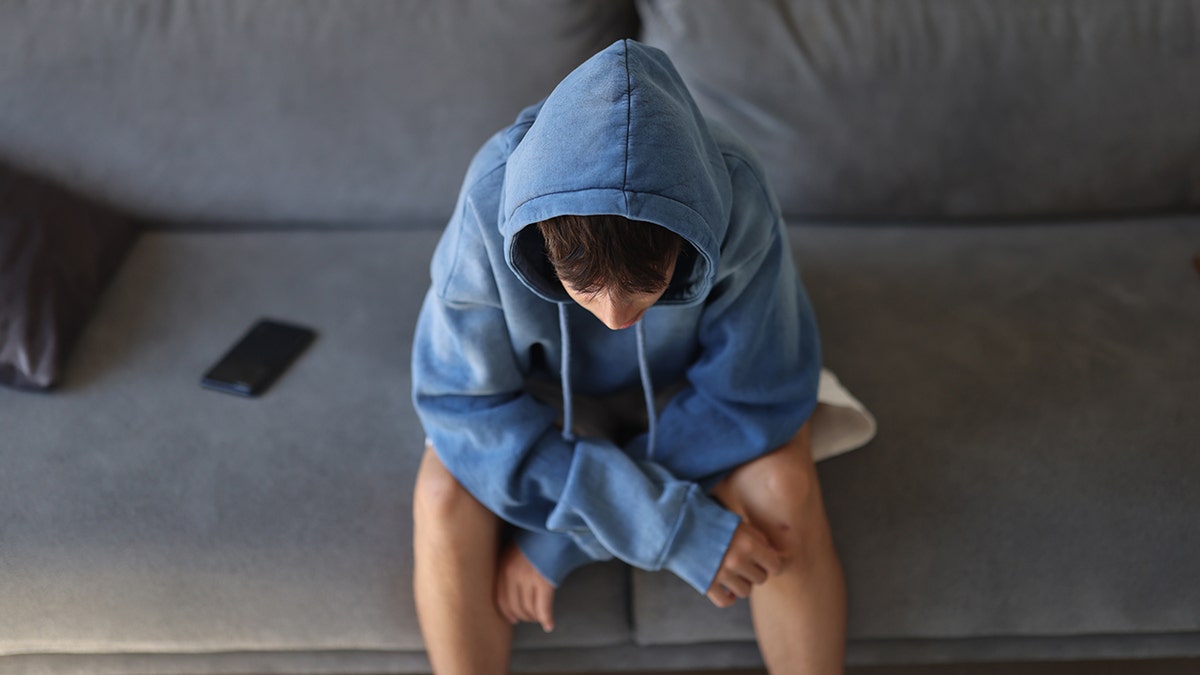 Young boy sits on couch in blue hoodie next to phone