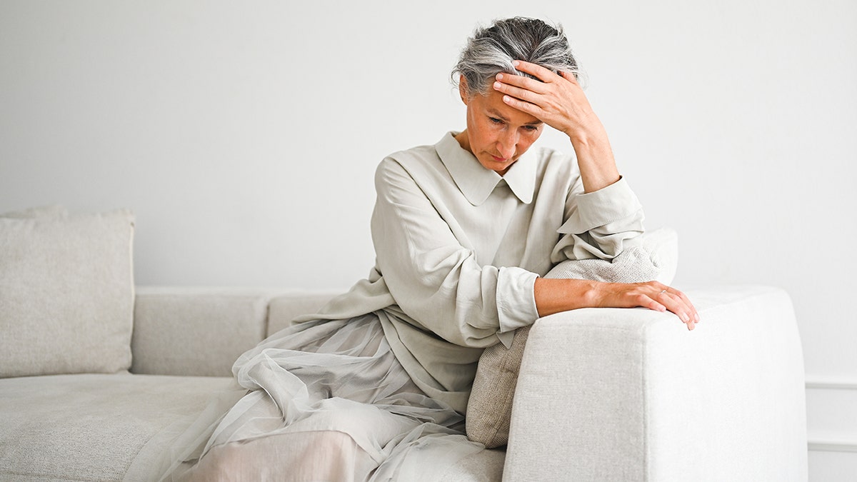 senior woman sits on white couch with her hand on her forehead