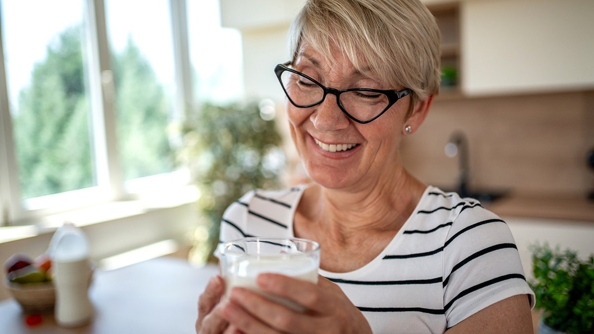 Smiling senior woman holding a glass of milk while sitting at a kitchen table, savoring a healthy breakfast and enjoying moments of comfort and relaxation in her home