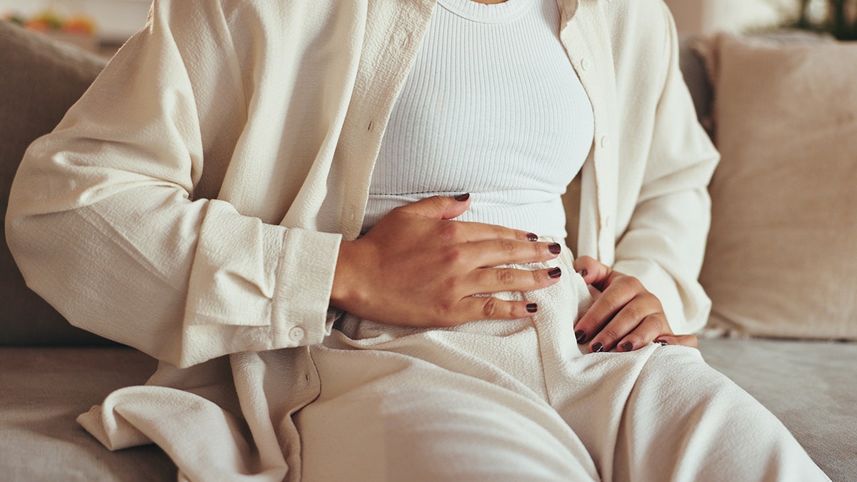 Woman seen sitting on coach holding abdomen area indicating pain or discomfort.